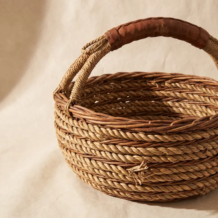 A large coiled willow gathering basket with a worn handle on a warm paper sweep — upper detail.
