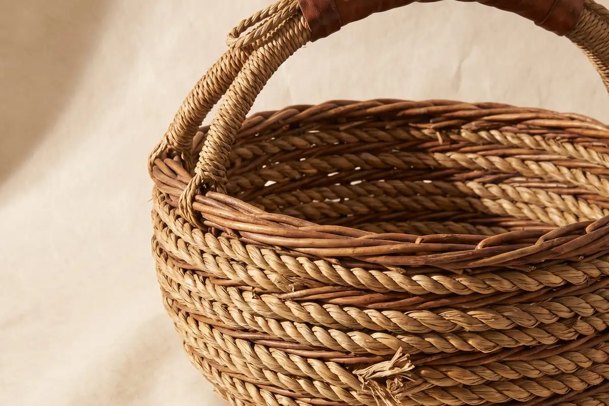 A large coiled willow gathering basket with a worn handle on a warm paper sweep — full width on the paper sweep.