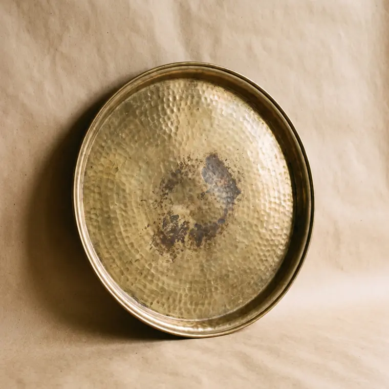 A close view of a small hammered brass dish with warm patina on a warm paper sweep — lower detail, showing the foot and the shadow.