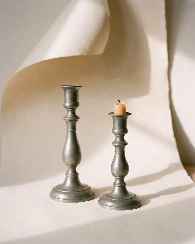 A pair of tall turned pewter candlesticks with soft grey patina at different heights on a warm paper sweep.