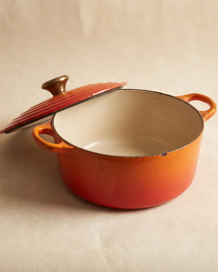 A vintage flame-orange enamelled cast iron casserole with a cream interior and a brass knob on a warm paper sweep.