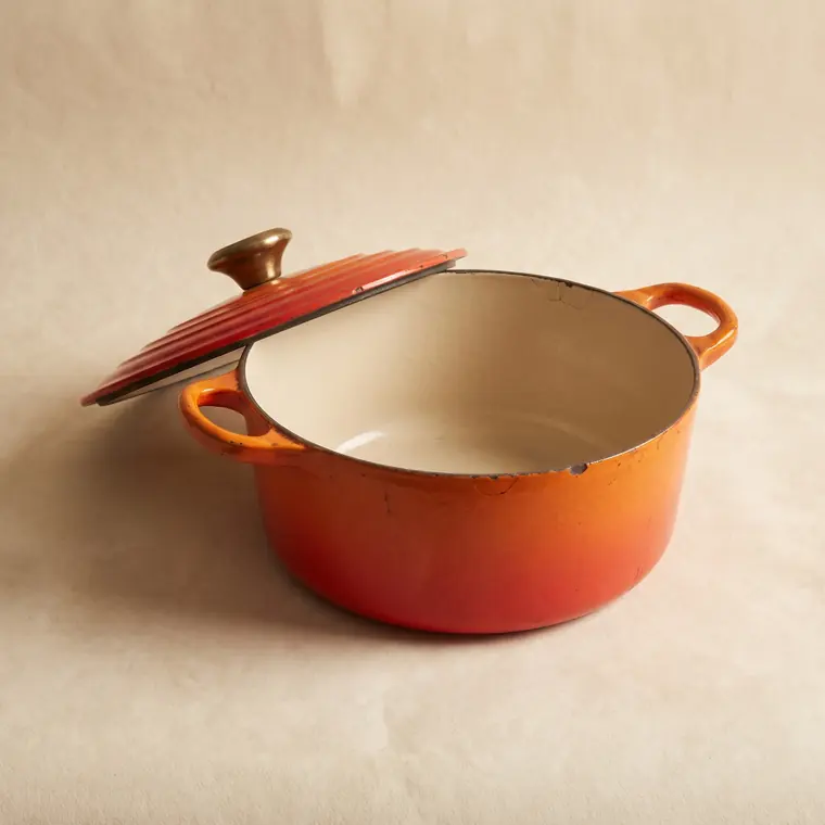 A vintage flame-orange enamelled cast iron casserole with a cream interior and a brass knob on a warm paper sweep — lower detail, showing the foot and the shadow.