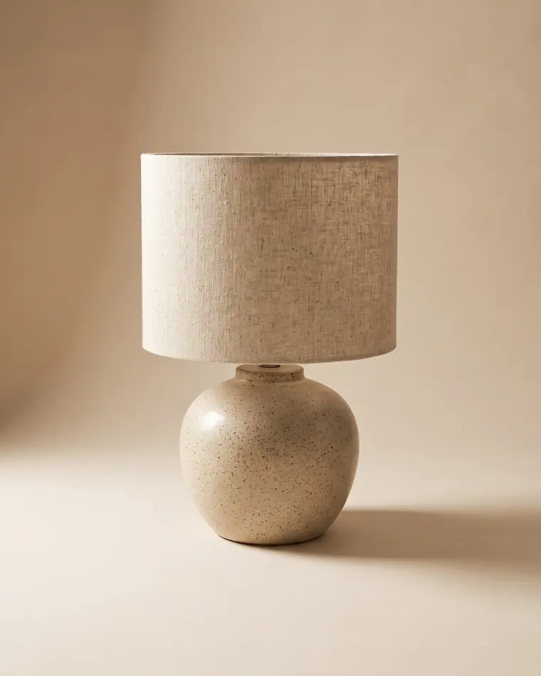 A ceramic table lamp with a fat speckled oatmeal stoneware base and a natural raw linen drum shade on a warm paper sweep.