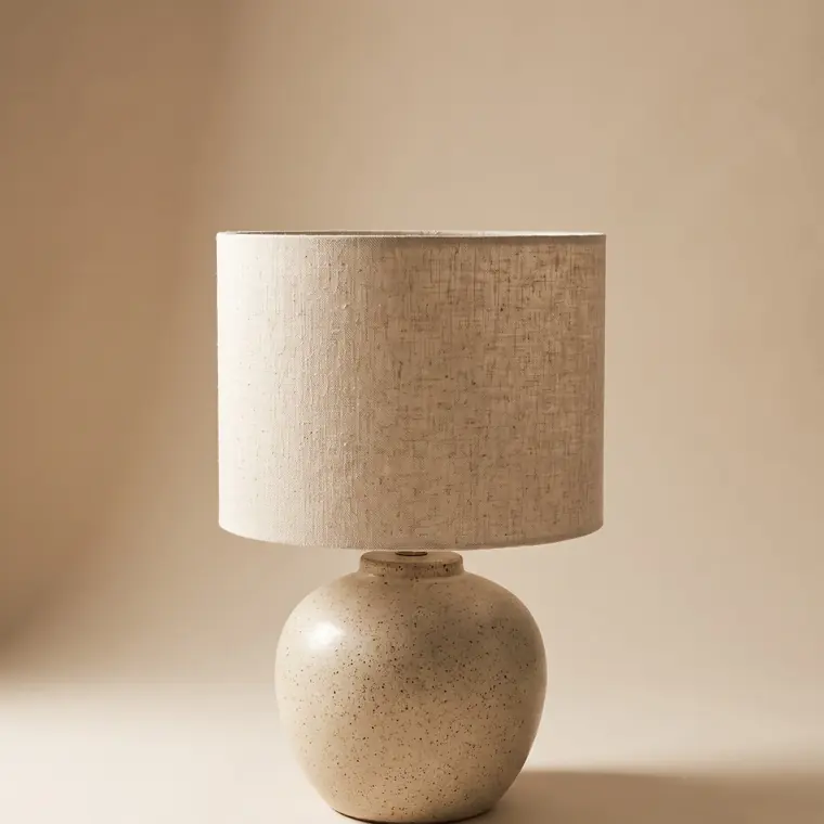 A ceramic table lamp with a fat speckled oatmeal stoneware base and a natural raw linen drum shade on a warm paper sweep — upper detail.