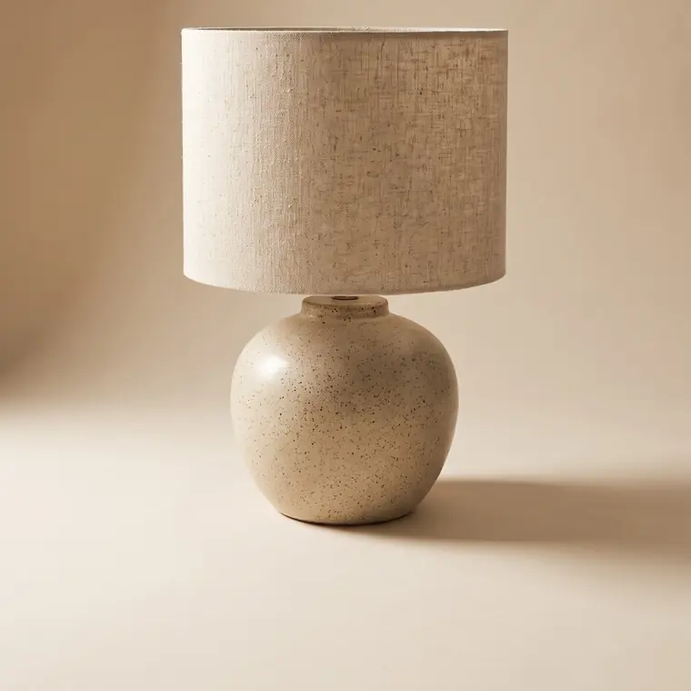 A ceramic table lamp with a fat speckled oatmeal stoneware base and a natural raw linen drum shade on a warm paper sweep — lower detail, showing the foot and the shadow.