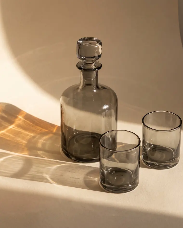 A hand-blown smoke grey glass decanter with a heavy stopper and two matching tumblers on a warm paper sweep.