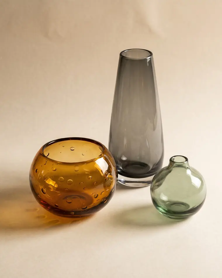 Three hand-blown studio glass vessels in smoke grey, amber and sage, casting warm coloured light on a warm paper sweep.