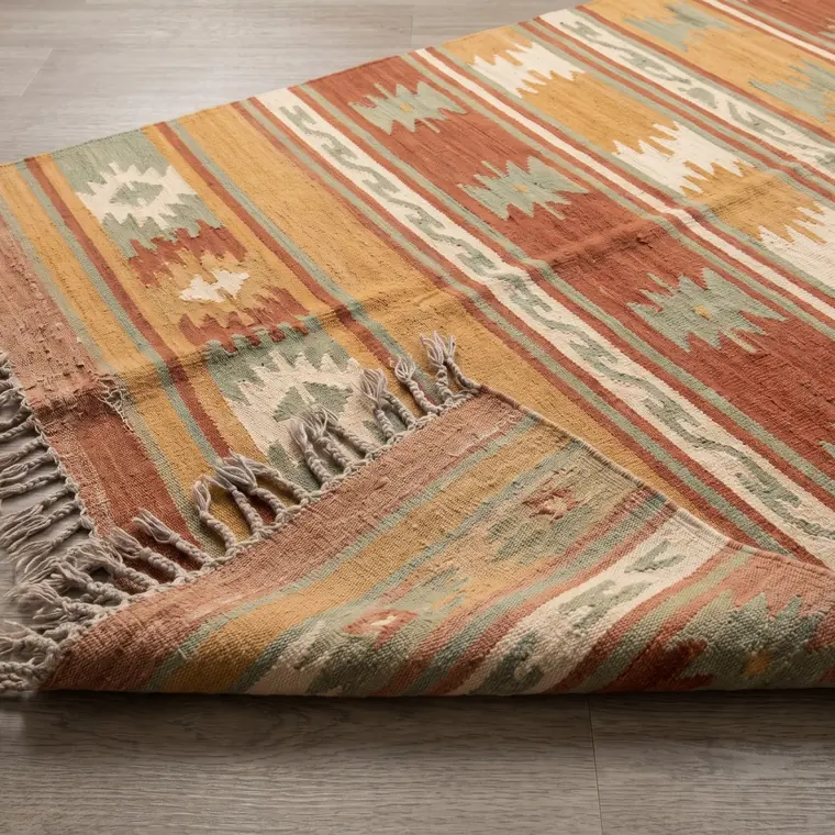 A worn handwoven wool flatweave rug in muted ochre and rust photographed from above — lower detail, showing the foot and the shadow.