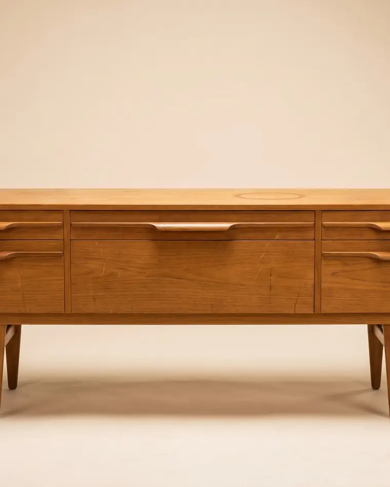 A long low Danish teak sideboard with sculpted drawer pulls and tapered legs against a warm paper backdrop.