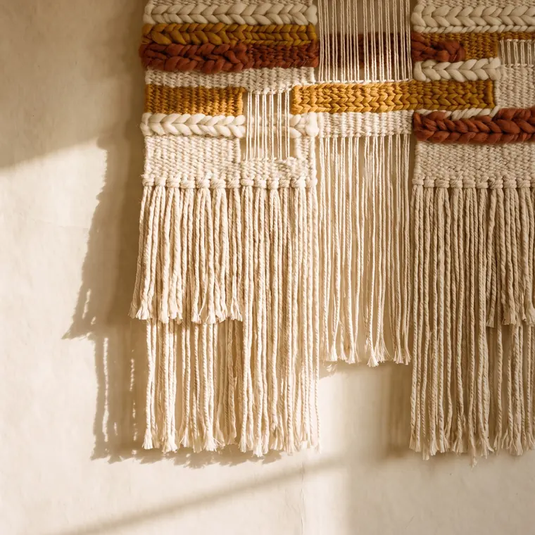 A close view of a small handwoven wool hanging with knotted fringe on an oak dowel — lower detail, showing the foot and the shadow.