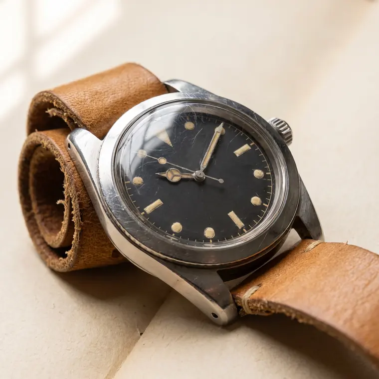 A close view of a vintage steel skin-diver watch with a browned tropical dial on warm paper — lower detail, showing the foot and the shadow.